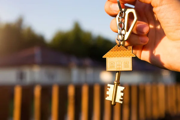 Key ring with keychain house in hand. Background of fence and cottage. Moving to a new home, mortgage, buying real estate, renting and booking housing, dream of living  in the village. Copy space