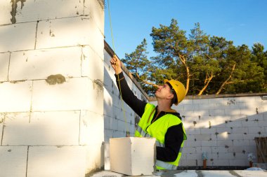 İnşaat işçisi, pencere açıklığının uzunluğunu ve duvarın uzunluğunu mezurayla ölçüyor. Kulübe gözenekli beton bloklar, koruyucu giysiler - kalın şapka ve bir yelek