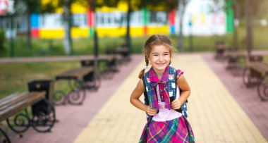 Okul bahçesinde okul üniformalı, sırt çantalı neşeli bir kız. 1 Eylül 'de okula dönüyorum. Mutlu bir öğrenci. İlköğretim, ilkokul. Bir öğrencinin portresi