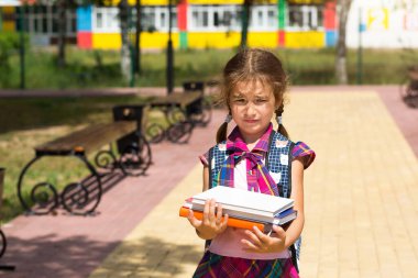 Girl with a backpack and a stack of books near the school. Back to school, the child is tired, heavy textbooks. Education, primary school classes, the beginning of the school year, September 1 clipart