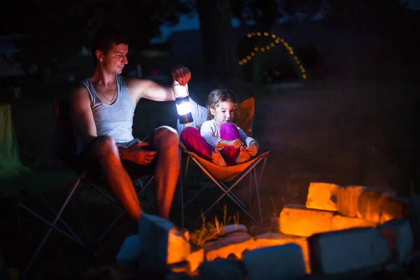 Dad and daughter sit at night by the fire in the open air in the summer in nature. Family camping trip, gatherings around the campfire. Father\'s Day, barbecue. Camping lantern and tent