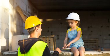 Dad and daughter in hardhat are at construction site of their future home. Choice of future profession of builder is inherited by child. The expectation of moving, the dream of a house. Mortgage, loan clipart
