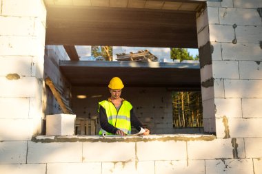 Sarı kasketli bir inşaat mühendisi ve tasarımcı inşaat alanındaki bir binanın çizimini inceliyor. Gözenekli beton bloklardan yapılmış bir ev, pencere ve duvarda bir işçi