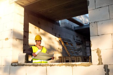Sarı kasketli bir inşaat mühendisi ve tasarımcı inşaat alanındaki bir binanın çizimini inceliyor. Gözenekli beton bloklardan yapılmış bir ev, pencere ve duvarda bir işçi