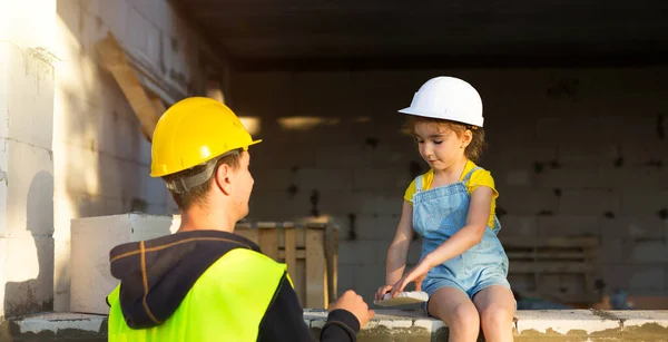 Dad and daughter in hardhat are at construction site of their future home. Choice of future profession of builder is inherited by child. The expectation of moving, the dream of a house. Mortgage, loan