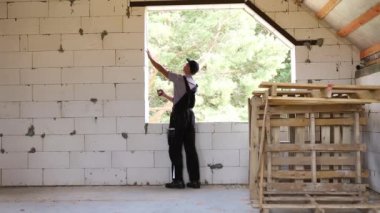 İnşaat işçisi, pencere açıklığının uzunluğunu ve tuğla duvarın uzunluğunu mezurayla ölçüyor. Kulübe gözenekli beton bloklar, koruyucu giysiler - tulum ve yelek