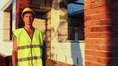 Proje mimarı, duvarları gözenekli beton blok ve tuğladan yapılmış bir evin inşaat sahasının yanında duruyor. Sarı baret ve yansıtıcı yelek giyen inşaat işçisi..
