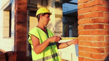 İnşaat işçisi, pencere açıklığının uzunluğunu ve tuğla duvarın uzunluğunu mezurayla ölçüyor. Kulübe gözenekli beton bloklar, koruyucu giysiler - kalın şapka ve yelek