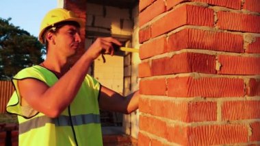 İnşaat işçisi, pencere açıklığının uzunluğunu ve tuğla duvarın uzunluğunu mezurayla ölçüyor. Kulübe gözenekli beton bloklar, koruyucu giysiler - kalın şapka ve yelek