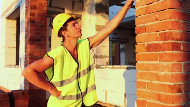 项目建筑师站在有墙的房屋的建筑工地附近 由多孔混凝土砌块和砖块组成 戴着黄色防护帽和反光背心的建筑工人 — 图库视频影像