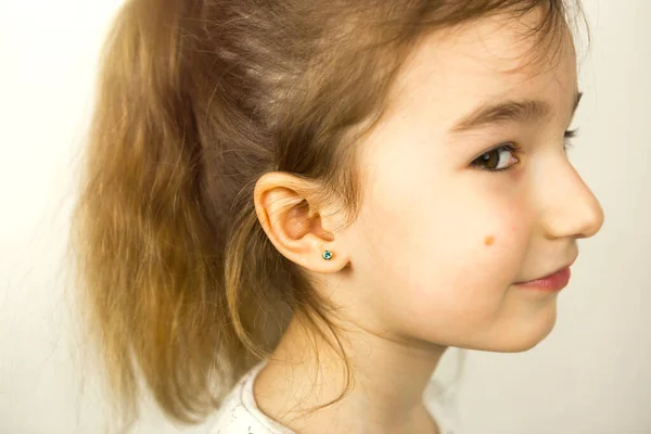 Ear piercing in a child - a girl shows an earring in her ear made of a medical alloy. White background, portrait of a girl with a mole on his cheek in profile.