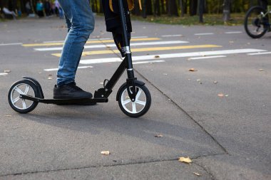 Sonbaharda parktaki scooter 'da kot pantolon ve spor ayakkabı giyen ve asfaltta kurumuş sarı yaprakları olan bir adamın bacakları. Sonbahar yürüyüşleri, aktif yaşam tarzı, çevre dostu ulaşım, trafik