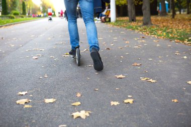 Sonbaharda parktaki scooter 'da kot pantolon ve spor ayakkabı giyen ve asfaltta kurumuş sarı yaprakları olan bir adamın bacakları. Sonbahar yürüyüşleri, aktif yaşam tarzı, çevre dostu ulaşım, trafik