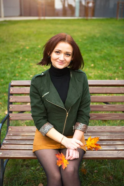 A woman with a bob haircut and a yellow fallen maple leaf in a green jacket and a mustard mini skirt. Autumn portrait on a park bench in a closed position, leg on leg. Autumn mood and fashion