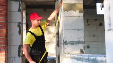 İnşaat işçisi, pencere açıklığının uzunluğunu ve tuğla duvarın uzunluğunu mezurayla ölçüyor. Kulübe gözenekli beton bloklar, iş kıyafetleri - tulum ve beyzbol şapkasından yapılmıştır.