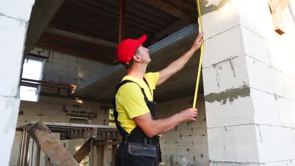 建設現場の建設労働者は テープ測定で窓の開口部とレンガ壁の長さを測定します コテージは多孔質コンクリートブロック 作業服で作られています ジャンプスーツと野球帽 — ストック動画