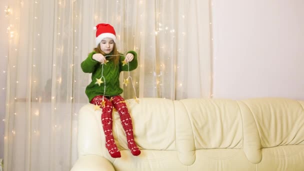 Dziewczyna Kapeluszu Mikołaja Ciepłym Swetrze Odkrywa Girlandę Gwiazd Domu Kanapie — Wideo stockowe
