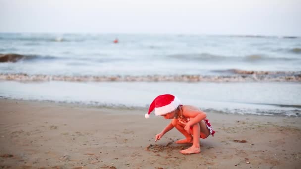 5岁的女孩穿着红色泳衣 头戴圣诞礼帽 在沙滩上玩耍 圣诞和新年到温暖的国家旅游 热带假日旅游 — 图库视频影像