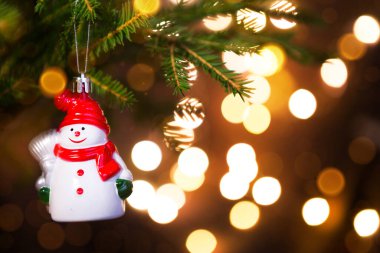 Kırmızı şapkalı Noel oyuncağı Kardan Adam, köknar ağacının dalında canlı bir eşarpla defocus 'ta altın renkli çelenk ışıkları. Yeni yıl, Noel, tatil geçmişi, bokeh, kopyalama alanı