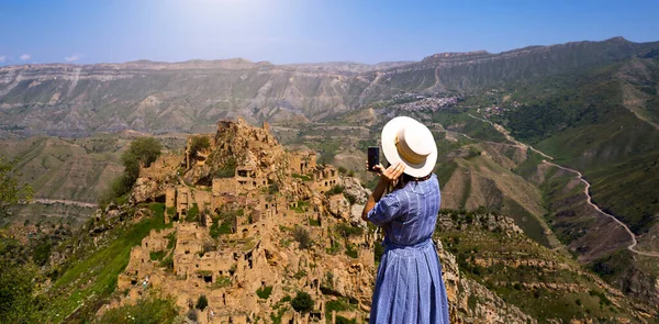 Dağıstan 'ın yüksek dağlarındaki kadın turist doğanın nefes kesen panoramik manzarasına ve terk edilmiş antik Gamsutl şehrine hayran. Çekim ve harabe fotoğrafları çekiyor, akıllı telefondan selfie çekiyor.