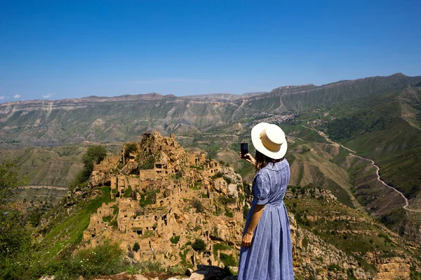 Dağıstan 'ın yüksek dağlarındaki kadın turist doğanın nefes kesen panoramik manzarasına ve terk edilmiş antik Gamsutl şehrine hayran. Çekim ve harabe fotoğrafları çekiyor, akıllı telefondan selfie çekiyor.