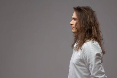 Stüdyoda uzun saçlı bir gencin profili. Beyaz gömlekli çekici adam gri arka planda izole edilmiş. Metnin için boşluk kopyala