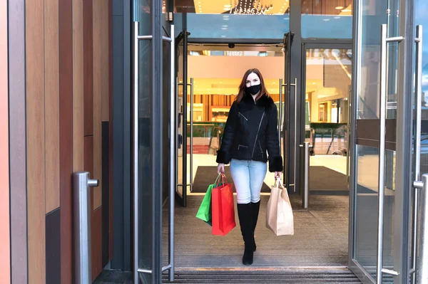 Beautiful woman in protective mask with shopping bags walks out mall. Stylish shopper girl