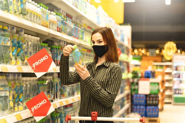 Süpermarkette maskeli bir kadın market alışverişi yapıyor. Müşteri hipermarkette ürün kompozisyonu okuyor. Kız içkiyi seçiyor. Mağazada müşteri var