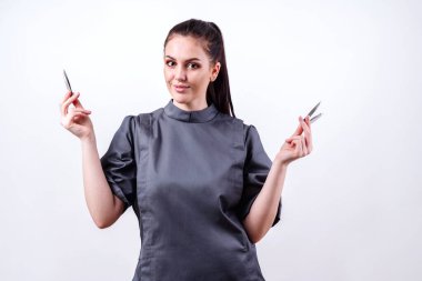 Esmer, ellerinde kaş cımbızı gösteriyor. Stüdyoda beyaz arka planda aletleri olan kız güzellik uzmanı.