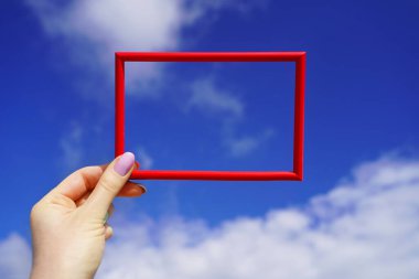 Mavi gökyüzüne karşı dişi elinde kırmızı dikdörtgen yatay çerçeve. Güneşli bir geçmiş. Metnin için parlak boşluk