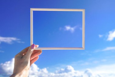 Mavi gökyüzüne karşı dişi elinde beyaz, boş, yatay bir çerçeve. Güneşli bir geçmiş. Metnin için boşluk