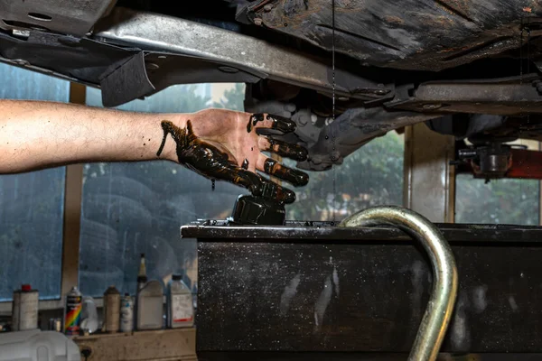 The car mechanic unscrews the diesel oil filter next to the oil pan, old black oil drains into the container.