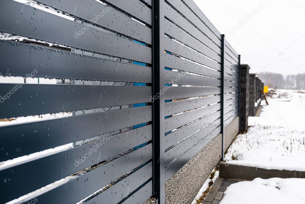 Modern anthracite panel fencing, with a visible fence foundation connector, it rains in winter, in the background there is snow lying on the ground.