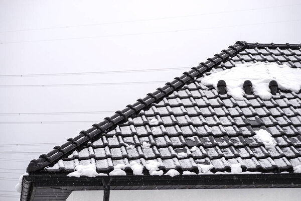 Крыша односемейного дома покрыта снегом на фоне облачного неба, видимый керамический вентиляционный камин на крыше и падающий снег.