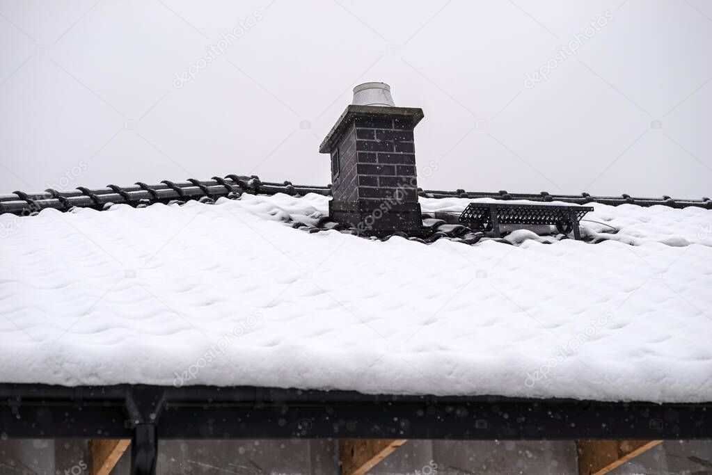 The roof of a detached house is covered with snow against a cloudy sky. Visible system chimney covered with tiles and falling snow.