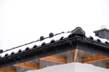 Tek bir aile evinin çatısı bulutlu bir gökyüzüne karşı karla kaplıdır. Görünür kiremitler, çatı kirişleri ve düşen karlar..