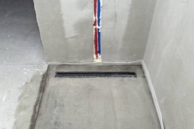 Duş zeminine kurulmuş modern siyah bir duş gideri, duvardaki görünür su boruları..