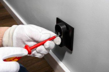 Elektrikçinin biri kırmızı tornavidayla siyah çift topraklı elektrik prizini oda duvarındaki elektrik kutusuna takıyor..