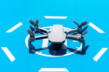 İHA mavi bir iniş sahasında duruyor, plastik pervaneler kapalı ve virajdaki kamera görülebilir durumda..