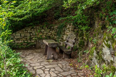 Eski bir bank ve ahşap bir masa, taş zeminde, ormandaki bir su kaynağının yanında, görülebilir taş bir duvar..