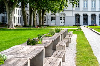 Parkta arka planda sıralanmış çiçek sıralı banklar ve masalar kemerli pencereli büyük bir bina..