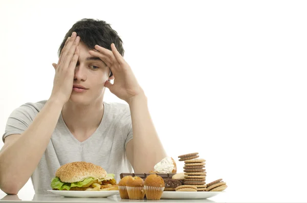 Młody człowiek z przodu trzyma mnóstwo ciasteczek i duży hamburger. wybór między czekolady, babeczki, ciastka i burger. Staramy się tłuszczu jedzenie fast food i dużą ilością cukru — Zdjęcie stockowe