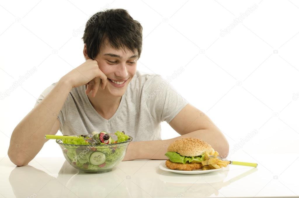 Young man holding in front a bowl of salad and a big hamburger. Choosing between good healthy food and bad unhealthy food. Organic food versus fast food