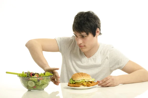 Giovane uomo che tiene davanti una ciotola di insalata e un hamburger grande. Scegliere tra buon cibo sano e cattivo cibo malsano. Cibo biologico contro fast food — Foto Stock