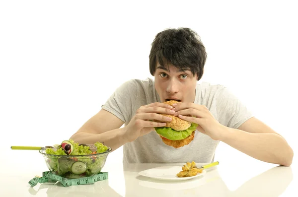 Giovane uomo che tiene davanti una ciotola di insalata e un hamburger grande. Scegliere tra buon cibo sano e cattivo cibo malsano. Cibo biologico contro fast food — Foto Stock