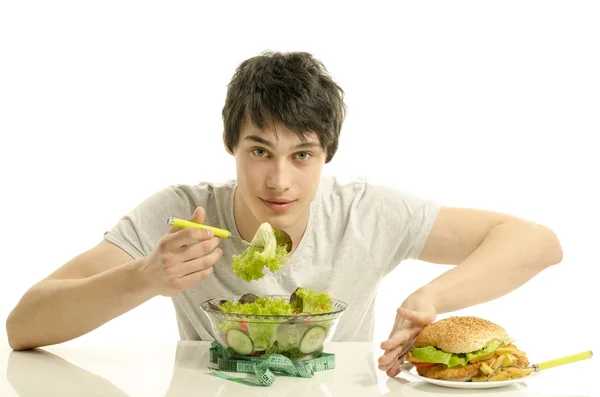 Young man holding in front a bowl of salad and a big hamburger. Choosing between good healthy food and bad unhealthy food. Organic food versus fast food — Stock Photo, Image