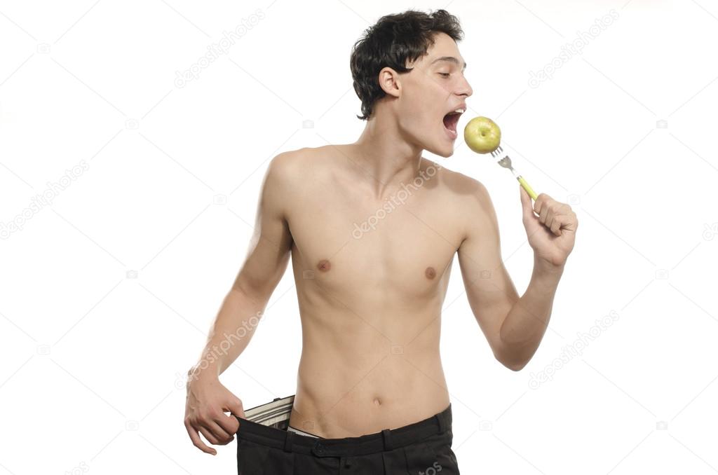 Skinny young man with large pants eating an apple,anorexic look, slim body. Man lost weight and comparing his larger pants from the days he was fat. Before and after, fat or slim