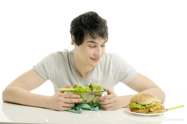 Giovane uomo che tiene davanti una ciotola di insalata e un hamburger grande. Scegliere tra buon cibo sano e cattivo cibo malsano. Cibo biologico contro fast food — Foto Stock