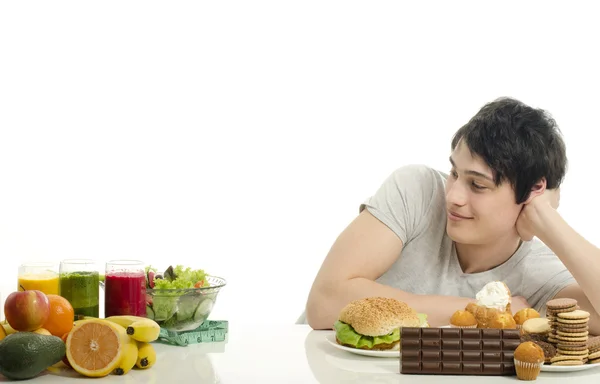 Hombre eligiendo entre frutas, batidos y alimentos orgánicos saludables contra dulces, azúcar, un montón de dulces y una gran hamburguesa, comida rápida —  Fotos de Stock