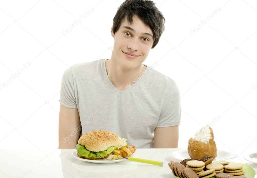 Young man holding in front lots of cookies and a big hamburger. Choosing between chocolate, cupcakes, biscuits and a burger. Trying to get fat eating fast food and lots of sugar